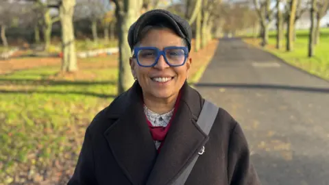 A woman wearing a brown coat smiles at the camera in a park 