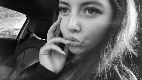 Cancer Research UK handout Black and white selfie of Sophie Claxton with long brown hair sitting in a car which her hand on her face and one nail resting on her lips. 
