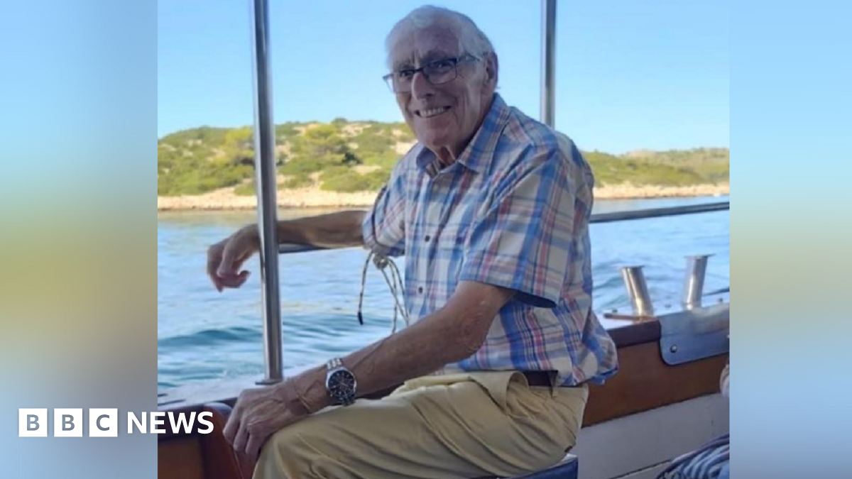 John Pryce Jones wears a blue checked shirt and beige trousers. He is on a boat. There is land in the distance and water in between