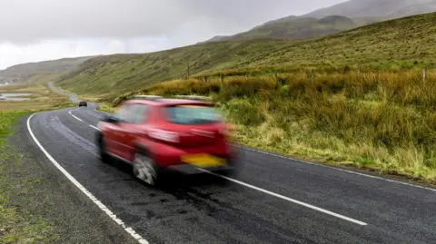 Getty Images A blurry red car driving on a single carriageway road in amongst hills and green grass
