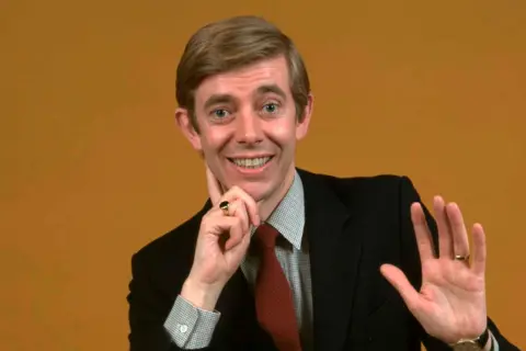 Getty Images Henry Kelly is wearing a dark suit jacket over a light-coloured dress shirt paired with a textured red tie. He is holding one hand near the chin, with a ring visible on the finger, while the other hand is raised slightly, also showing a ring and a wristwatch. The background is a solid mustard yellow colour, creating a clean and minimalistic setting.