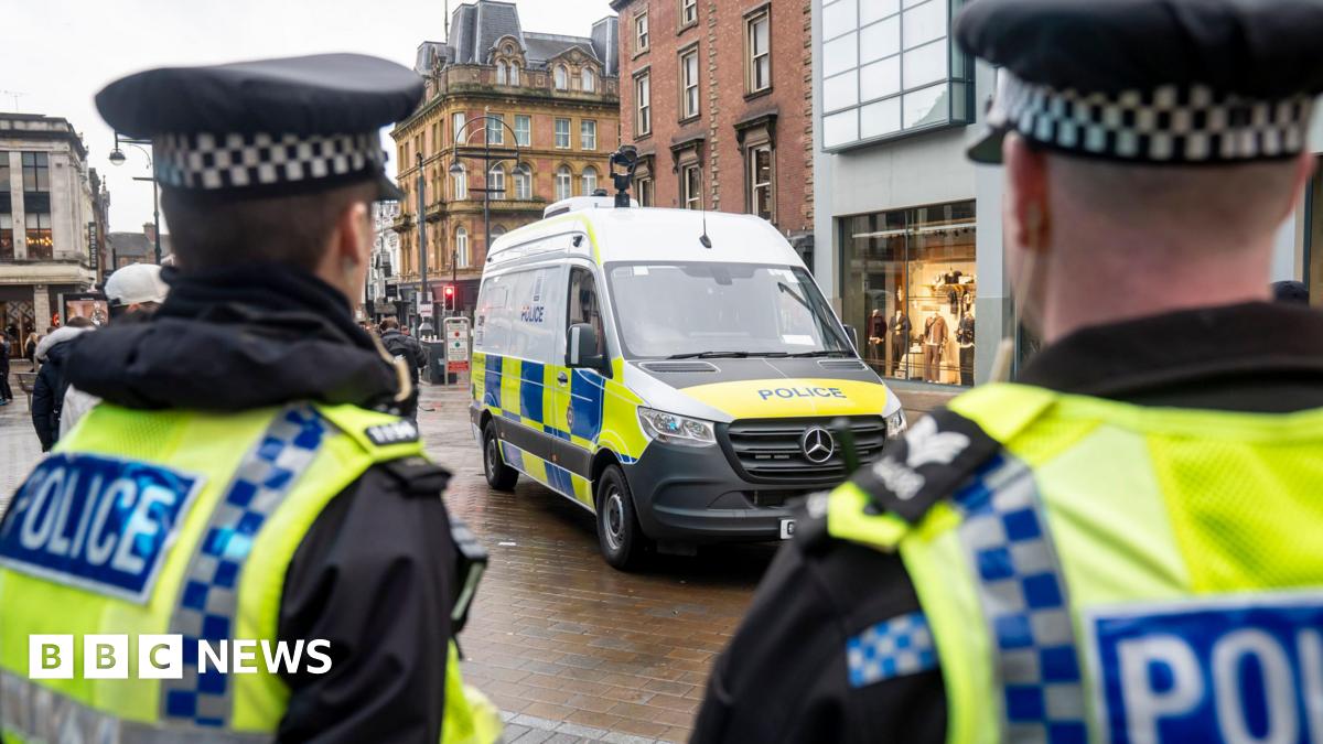 Two police officers stand with their backs to the camera, looking down Briggate in Leeds. They are wearing high-vis vests. They are looking at a police van, which is parked next to a shop.