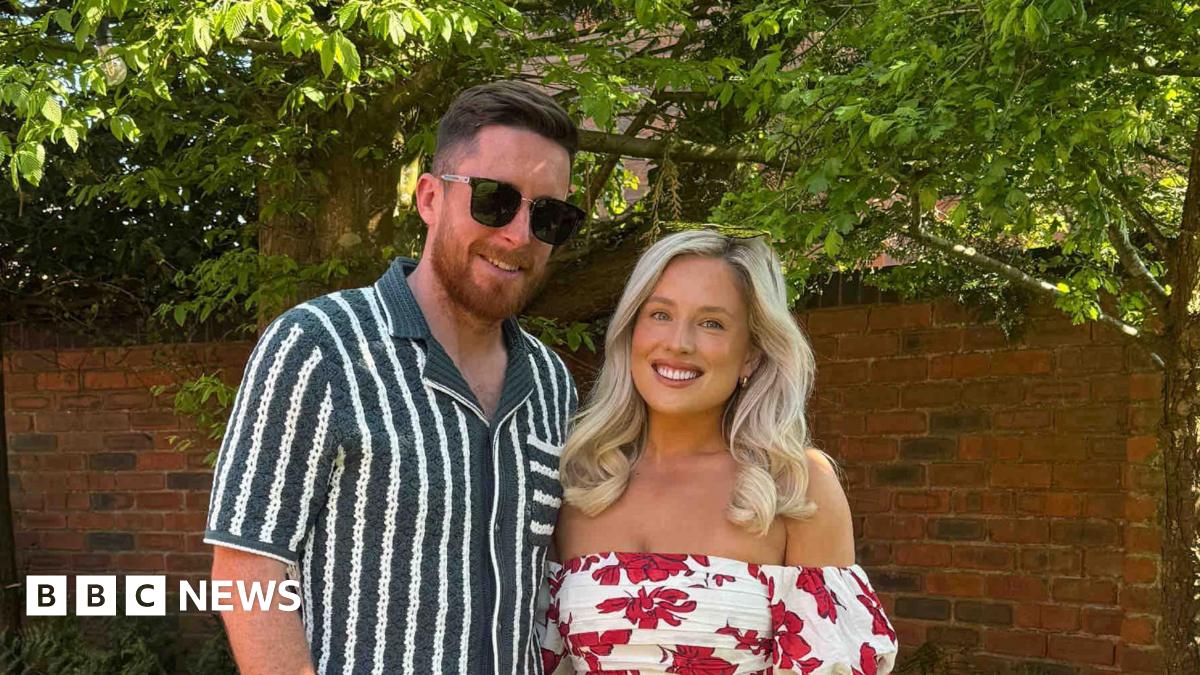Two people, Kat and Adam standing smiling in a garden. She wears a summer flowery dress and has blonde hair. he has brown hair and a beard and wears white trousers, a stripy shirt and sunglasses.