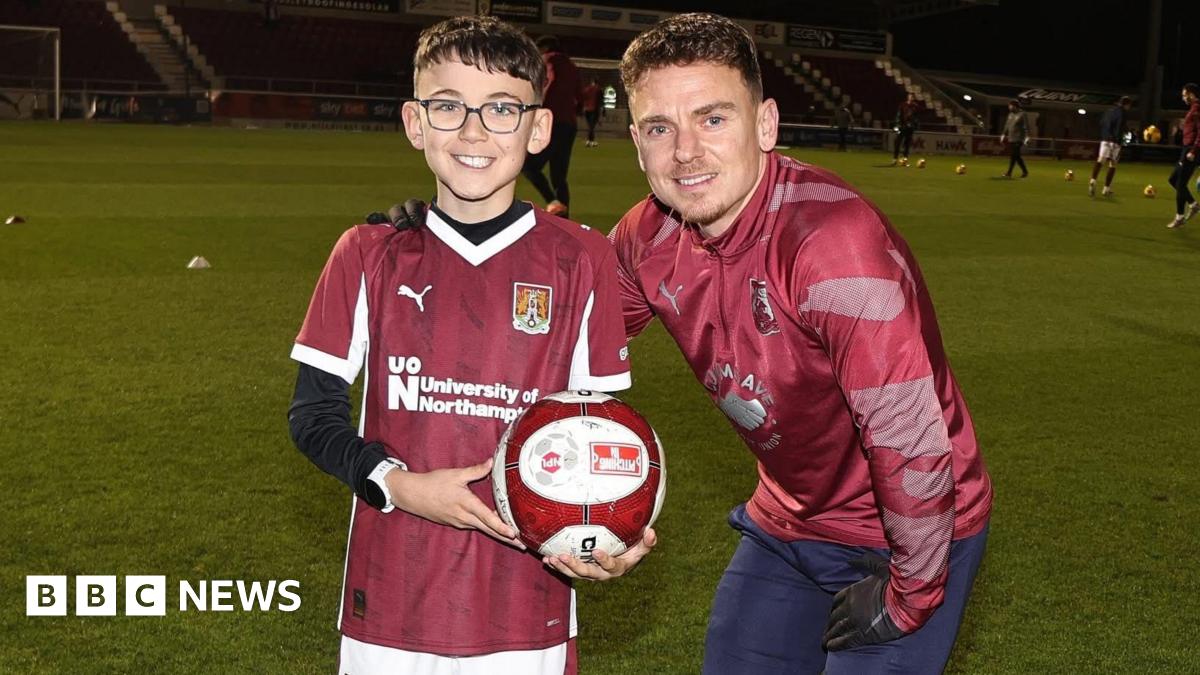 Ezra stands on the pitch next to Sam, who has his arm around Ezra's shoulder, and they both smile at the camera. Ezra holds the football which is in team colours of Maroon and white. He wears the team T-shirt with a University of Northampton logo on, over a black long-sleeved top. The pitch is lit up and there are some players behind them warming up.