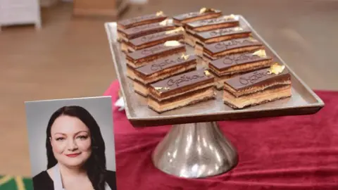 STVR A silver pedestal tray displays neatly arranged layered chocolate pastries with glossy tops, each decorated with the word “Opera” and a small piece of gold leaf. In front of the tray is a portrait photo of a woman wearing a light blue apron over a black top.