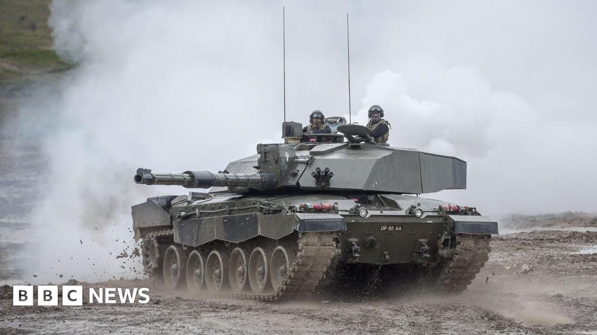 A tank pictured in Wiltshire in 2017. Two members of the armed forces drive through mud, with smoke in the background.