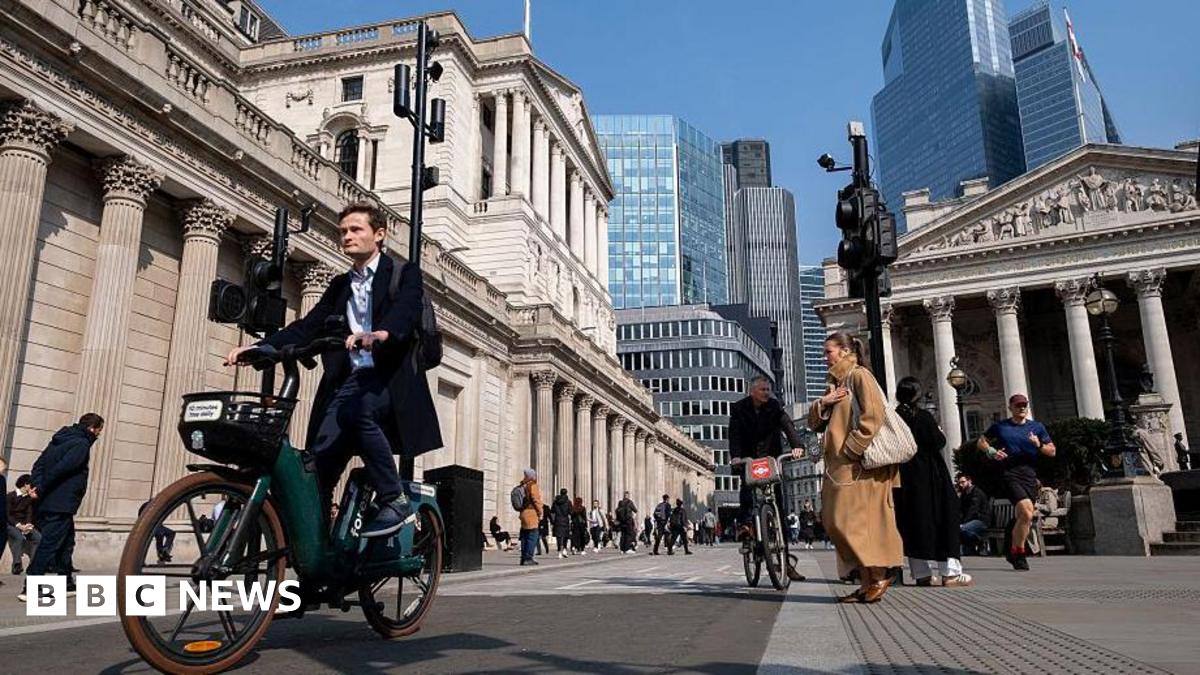 A man in a suit rides a bike in through the city in London with the Bank of England building behind him