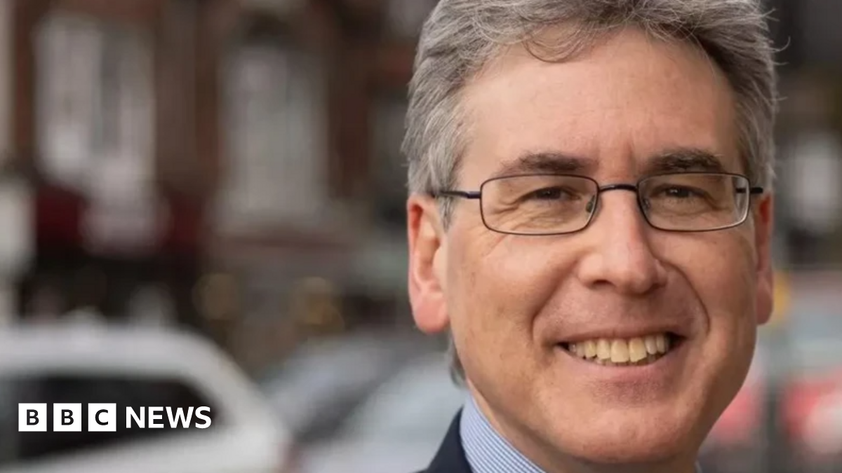 Mr Foster is smiling at the camera in a close-up photo. He has glasses on and a blue shirt and dark suit jacket. He is stood outside on a street.