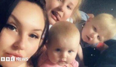 A young woman with dark hair alongside three blonde-haired toddlers. They are all looking directly into the camera.