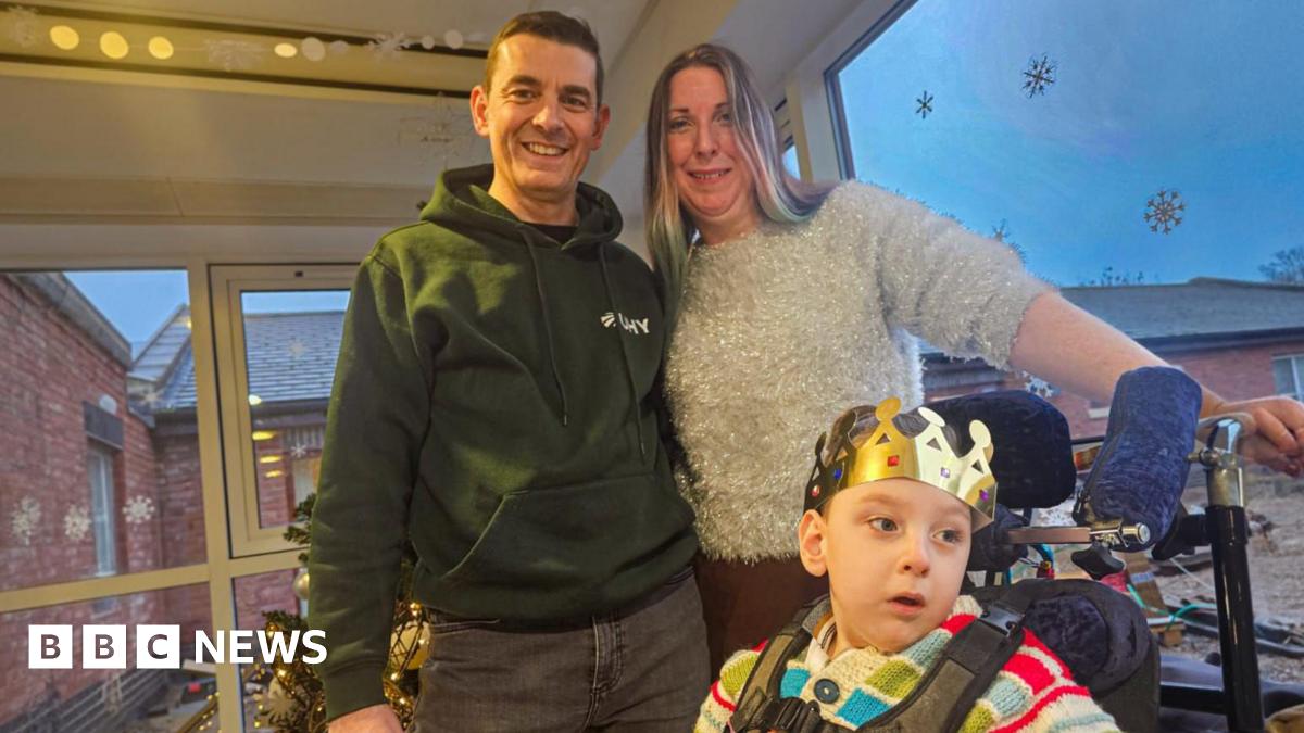 Dave and Emma stand with their arms around each other behind their son who is in a wheelchair. He has a gold grown on and it wearing a bright stripy jumper. Dave is wearing a green hoody and has dark brown hair and Emma is wearing a grey sparkly jumper and has blonde hair.