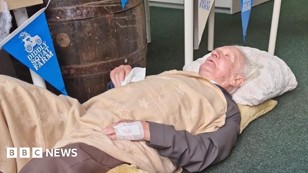 An elderly man lying on the floor of a garden centre, wrapped in blankets and with a pillow propping him up.