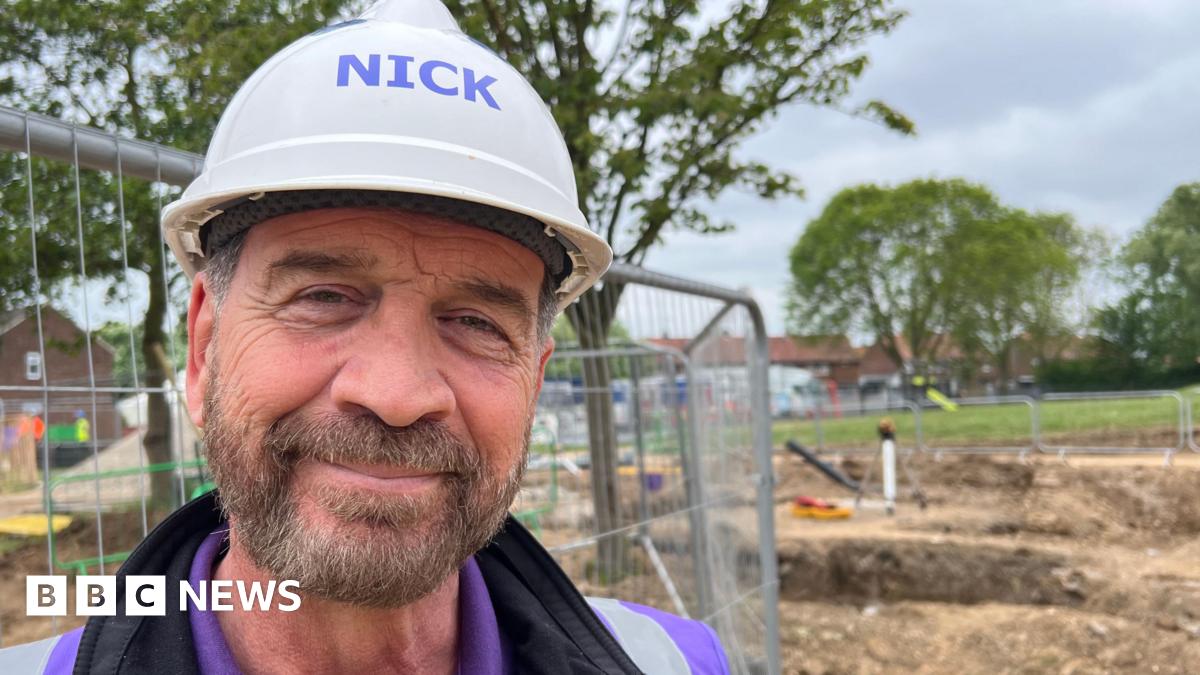 TV presenter Nick Knowles smiles into the camera. He has a beard and is wearing a white hard hat, purple t-shirt, black top and a purple high-vis vest. He is standing in front of a building site and a metal fence. Trees and houses are visible in the distance.