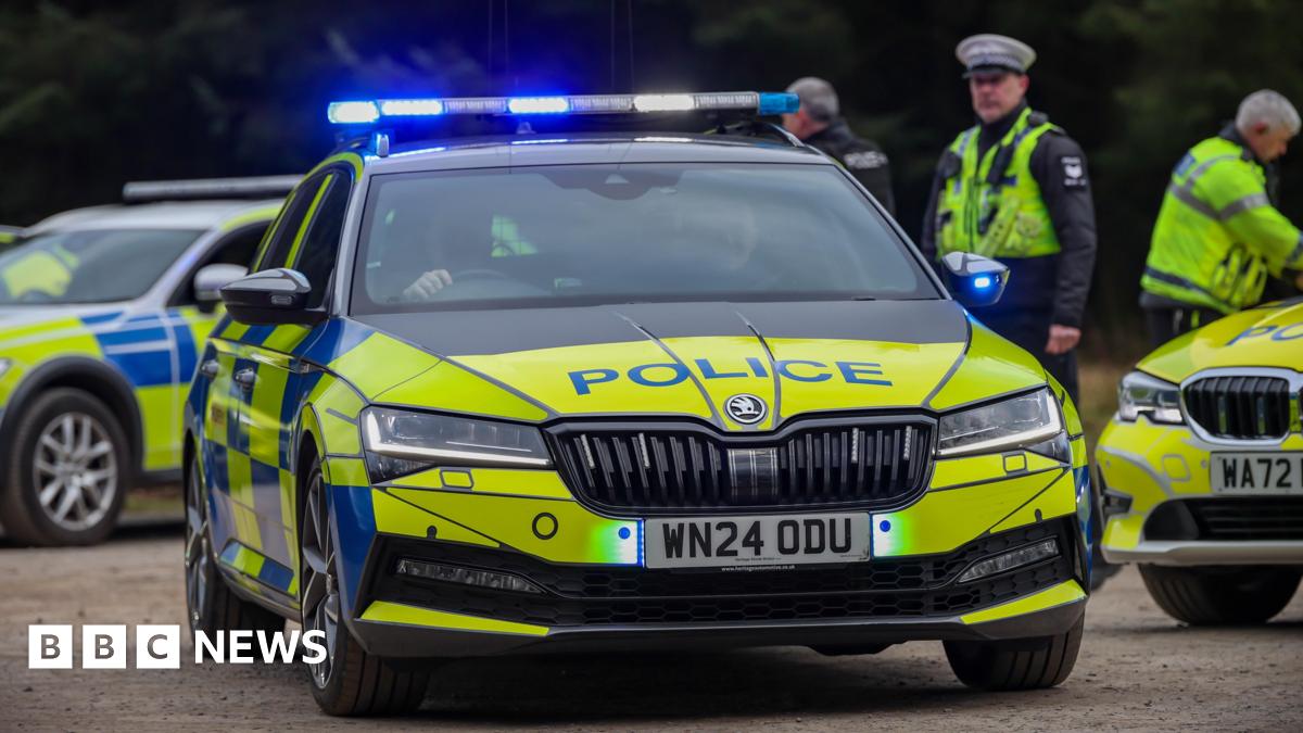 A Devon and Cornwall Police car with its blue lights on drives away from a car park. Two other police cars and three police officers are also in the car park. The car driving away is black, yellow and blue. It is a Skoda and has the licence plate WN24 0DU.