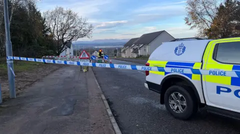 BBC Blue and white police tape stretches from a lamppost past a police truck that is parked in the middle of a road. The four-wheel drive truck is white with bright yellow and blue markings and a Police Scotland badge. Further down the road is a set of traffic lights at a roadworks and rows of houses.