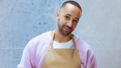 Channel 4/Love Productions A man with short black hair and beard stands with his head cocked to one side. He is wearing a purple cardigan over a white jumper, with a brown apron over the top.