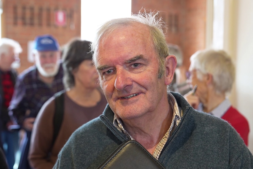 A retiree-aged man in a pullover smiles as he speaks to our reporter while people mill about.