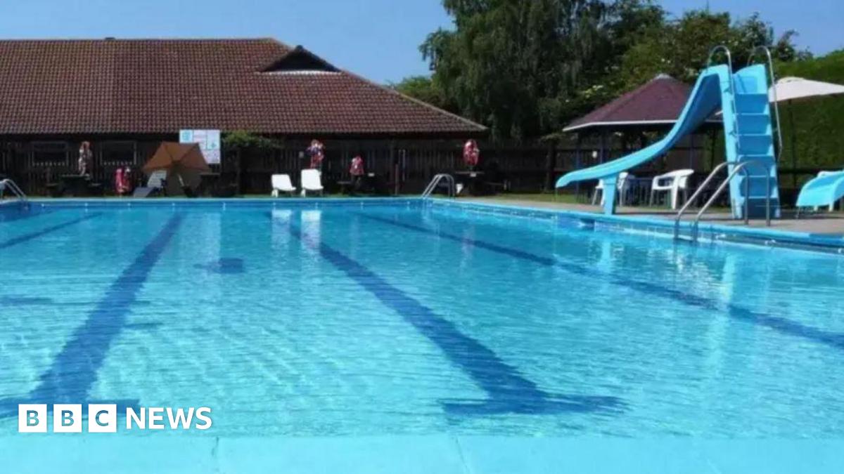 An outdoor swimming pool, with dark blue tiles at the bottom of the pool which mark out the lanes. To the right of the pool there is a blue slide. In the background are chalet-style buildings.