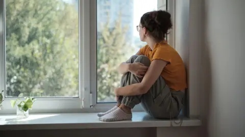 MomentoJpeg A sad upset teenager in a yellow t-shirt sitting on a windowsill hugging her knees looking out of a window
