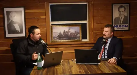 Contra Mundum Two men sit in front of microphones and computers, with patriotic artwork behind them, including a copy of a famous painting of George Washington during the US Revolutionary War and former presidents Richard Nixon and James Polk