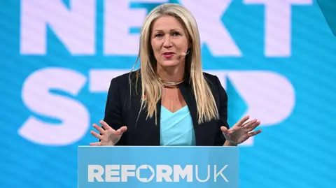 Getty Images Laura Anne Jones wearing a black jacket and blue top, standing at a podium with her hands raised. She has a white and blue backdrop behind her and is wearing a microphone.