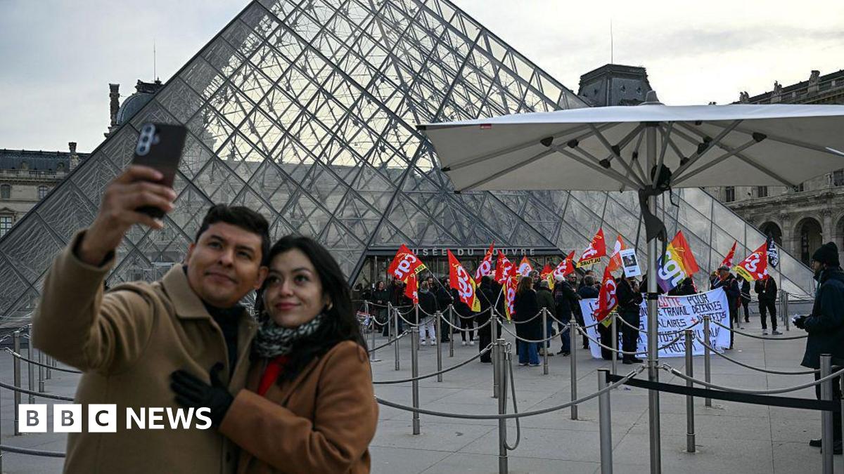 A couple take a selfie against the backdrop of strikers with placards at the Louvre Musuem