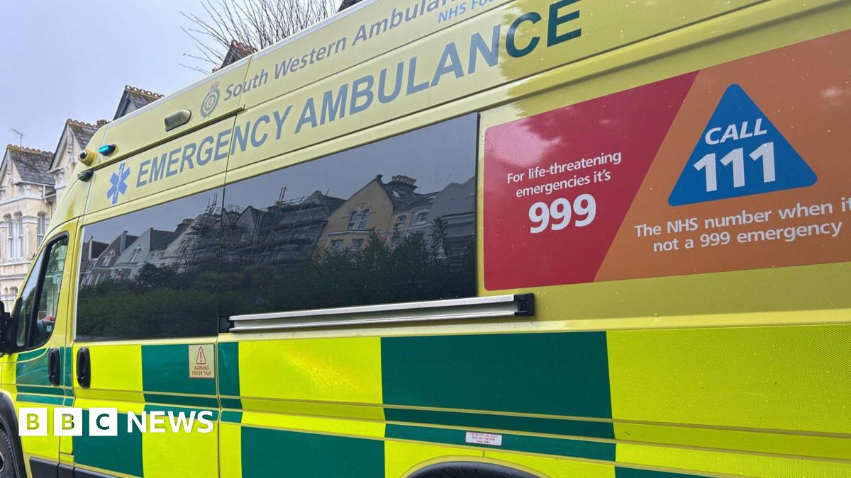 A side view of an ambulance belonging to the South Western Ambulance Service. It is bright yellow and green with a red and orange poster reading 'for life-threatening emergencies it's 999, call 111 the NHS number when it's not a 999 emergency. A few three-storey houses can be seen at the front of the ambulance.