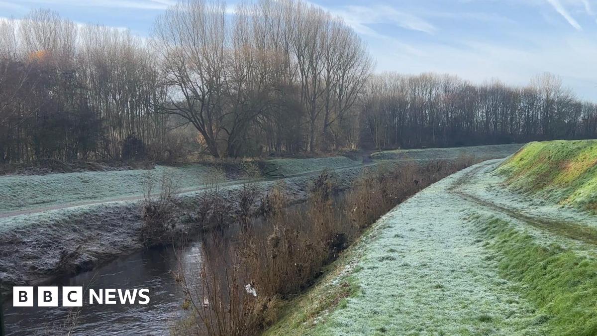 A green path covered by frost lines both sides of the River Mersey in Greater Manchester. Bare tree are on the left and bare shrubs line the river itself. It goes through a wider park.