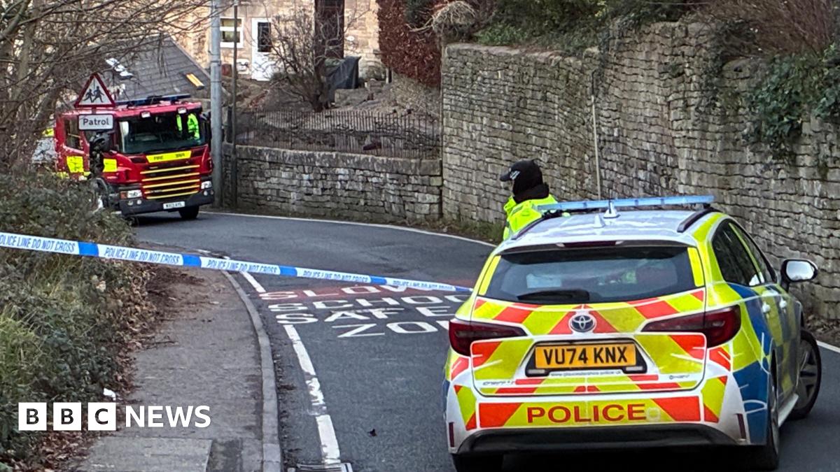 A police car blocks off a road in front of 'Police Do Not Cross tape' A fire engine is parked on the hill and a row of terrace properties can be made out behind a wall. One of the properties has been badly damaged by a fire, with its roof almost entirely missing with only the bare structure visible.