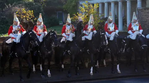 PA Media Men wearing ceremonial red suits hold swords sitting on hoses in front of a stone wall