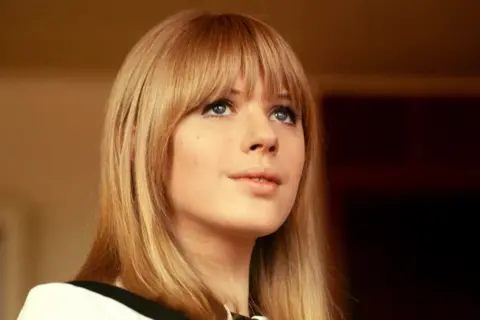 Getty Images Marianne Faithfull pictured in the 1960s. She has straight, shoulder-length light brown hair and blunt bangs, and is wearing a white top with a black collar detail. The background appears to be indoors with warm lighting and a dark area behind, suggesting an interior setting.
