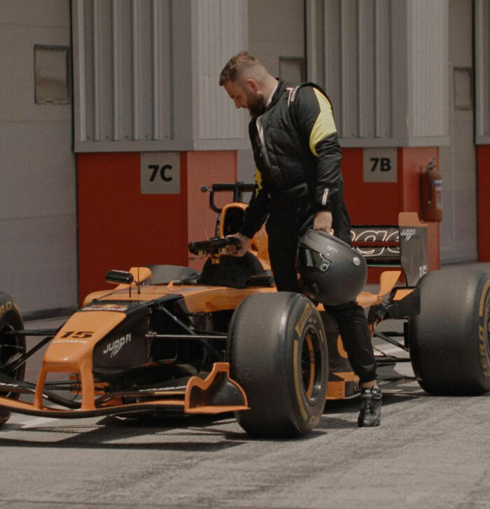 Man in racing suit holding helmet beside orange and black race car in pit area, unrelated to crypto scammer details.