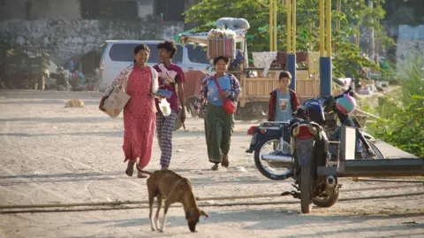 Lulu Luo/ BBC People walking down the street in Mandalay, past parked two-wheelers. Also pictured is a dog smelling the ground.