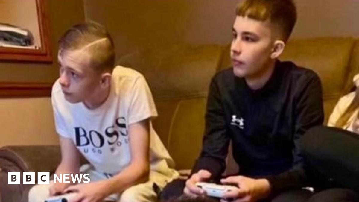 Mason Rist and Max Dixon are shown sitting on a sofa playing a video game together. Both hold controllers and look towards a screen out of shot, with their forearms leaning on their knees. Mason wears a white T-shirt while Max has a long-sleeved black top on.