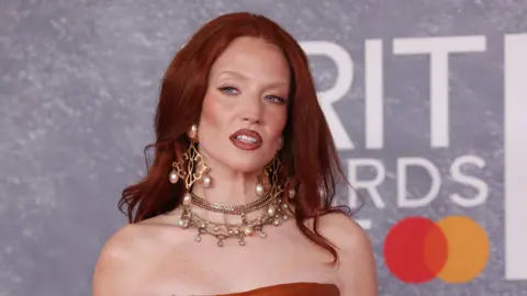 Getty Images Jess is wearing a strapless dark orange dress. She has on large gold and pearl dangly earrings. She has long ginger hair past her shoulders and blue eyes. 