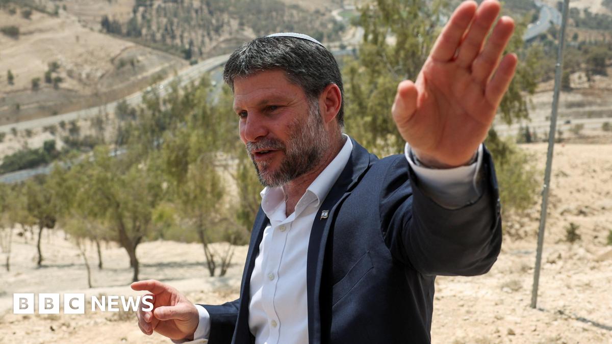 Israeli Finance Minister Bezalel Smotrich wears a white dress shirt and black suit jacket. He has short salt-and-pepper hair and beard. He is speaking and gesturing with his hands. Dry ground, shrubbery and a road winding through hills in the West Bank are seen behind him.