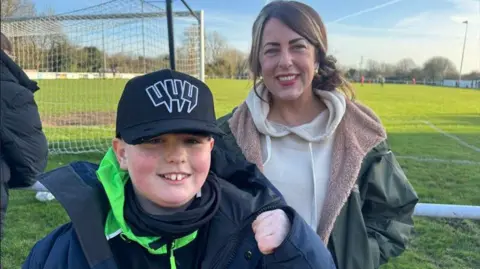 A young boy with a baseball cap, dark coloured tops and a blue coat is smiling while standing near a football pitch. He is standing next to a woman who is also smiling. She has brown hair tied back and is dressed in a warm fleece coat and hoodie.