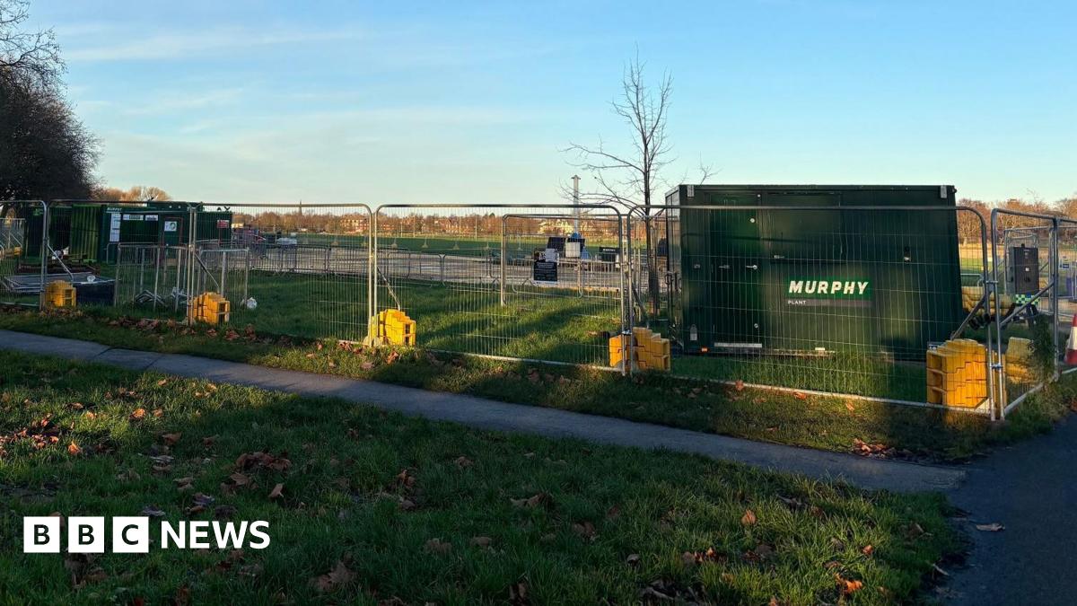 A large green tank stands at corner of green field surrounded by metal fences in a park.
