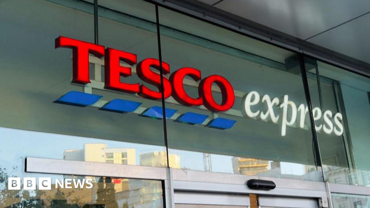 A Tesco Express sign, from a generic store, on glass above the entrance doors. "Tesco" is in red with blue dashes below it, and "express" is in white.