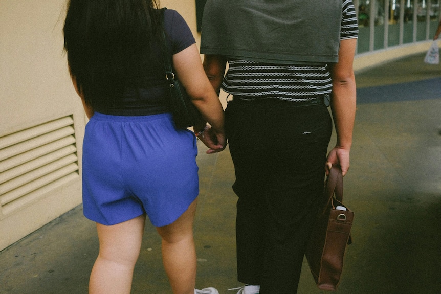 A woman in blue shorts and a man in a striped top walk hand in hand down a sidewalk.