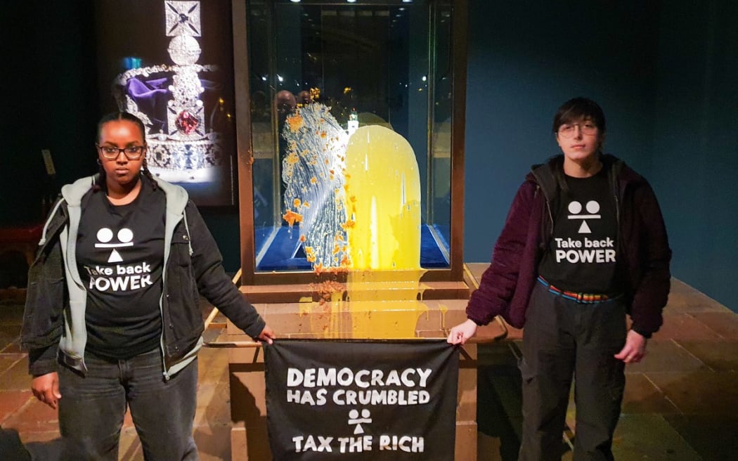 A handout photo taken on and released by Take Back Power on December 6, 2025, shows supporters of the civil resistance group holding a banner reading 'Democracy has crumbled - Tax The Rich' after they smothered dessert and custard over the case containing the Crown Jewels, at the Tower of London.