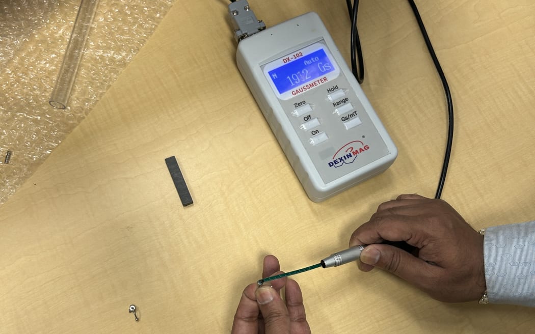 Photo showing a pair of hands holding a small magnet, and holding a machine to test it.