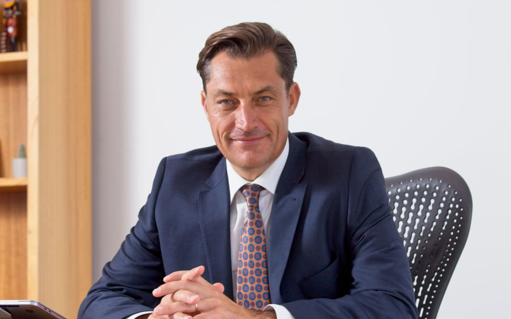 David is sat behind a desk. He wears a blue suit and looks towards the camera.