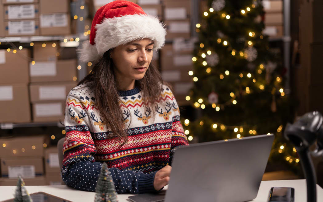 Salesperson in a Santa Claus hat takes an order by laptop computer while sitting in warehouse with boxes for Christmas. Shopping and delivery. Small business concept.
