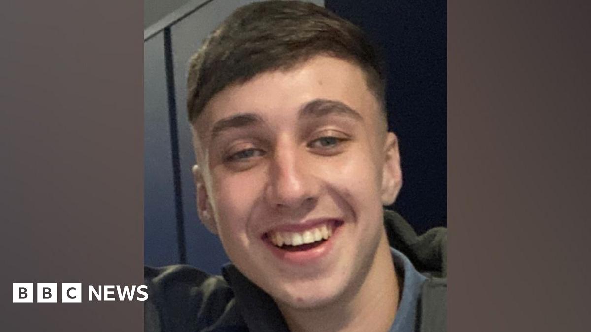 A close-up photograph of Jay Slater who is smiling widely. He has short, brown hair and is wearing a khaki-coloured jacket.