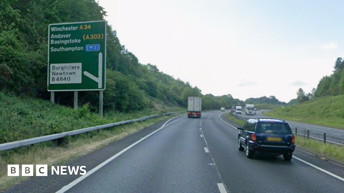 View of A34 southbound dual carriageway, a grey road with two lanes and a central reservation. There are green bushes to the left and a road sign which shows an upcoming exit for Burghclere. On the road is a black car with a blurred number plate and a lorry.