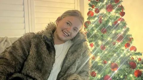 Jenna O'Donnell A woman with blonde hair in a fur jacket sitting in front of a Christmas tree with red baubles.
