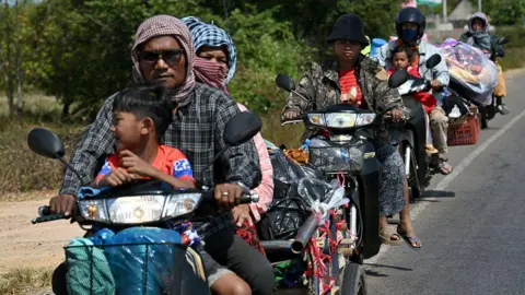 Getty Images A line of people riding down the side of a road on motorcycles, many of them carrying piles of possessions