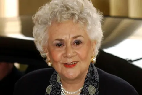 Getty Images Dame Joan Plowright has short, curly white hair and is wearing a dark outfit paired with a patterned scarf and a pearl necklace. Gold earrings are visible, and the background shows part of a vehicle and an indoor setting with warm lighting.
