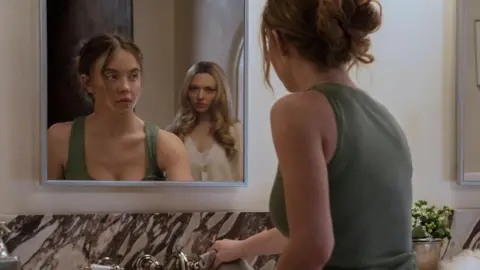 Lionsgate A scene from The Housemaid with Sydney Sweeney's reflection shown in the mirror. She is staring at Amanda Seyfried who appears behind her in the reflection. Sweeney is wearing a green vest and Seyfried is wearing a white blouse.