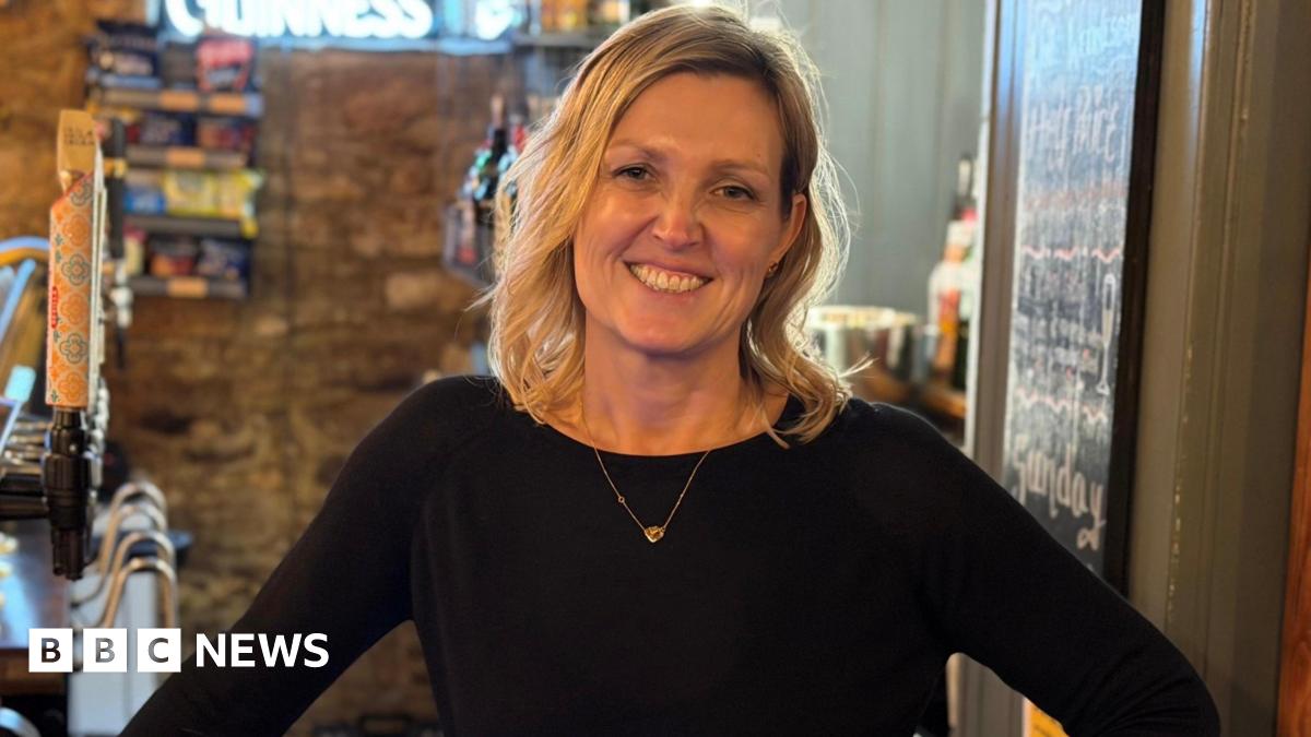 A blonde woman smiling at the camera. She is standing behind the bar in a pub. She is wearing a black top with a gold necklace.